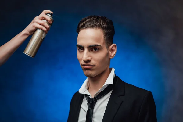 Hairdresser sprays the hair of a young elegantly dressed guy, preparing for photo shoot — Stock Photo, Image