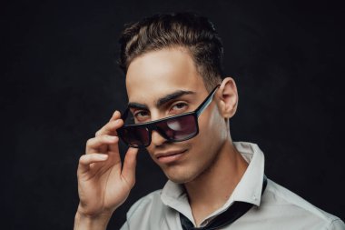 Beyaz gömlekli ve güneş gözlüklü yakışıklı ve kendinden emin bir genç. Kameraya karanlık bir arkaplanda bakıyor.