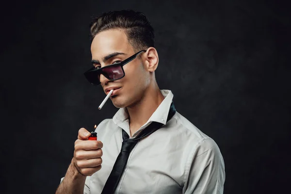 Joven hombre guapo en gafas de sol con camisa blanca y corbata negra enciende un cigarrillo —  Fotos de Stock