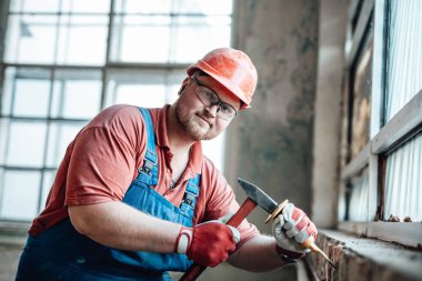 İnşaat alanındaki gülümseyen inşaatçı çekiçle tuğla bir duvarın üzerinde çalışıyor.