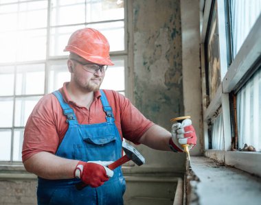 Gülümseyen profesyonel işçi pencere pervazından eski alçıyı temizliyor.