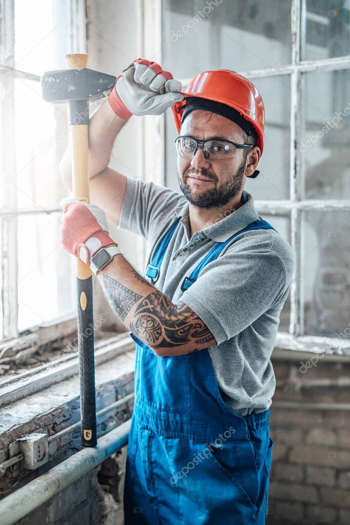 Muscular construction worker stands by the window and holds a big sledgehammer