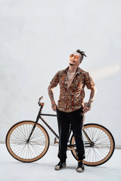 Stílusos tetovált gengszter szörny hawaii ingben és sportnadrágban, egy egyedi kerékpárral, őrült mosollyal a kamerába néz. — Stock Fotó