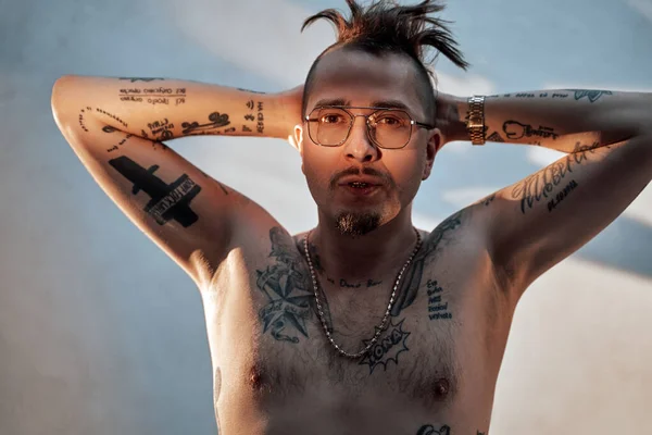 Elegante gángster en gafas con cuerpo desnudo tatuado y accesorios tomados de la mano detrás de su cabeza — Foto de Stock