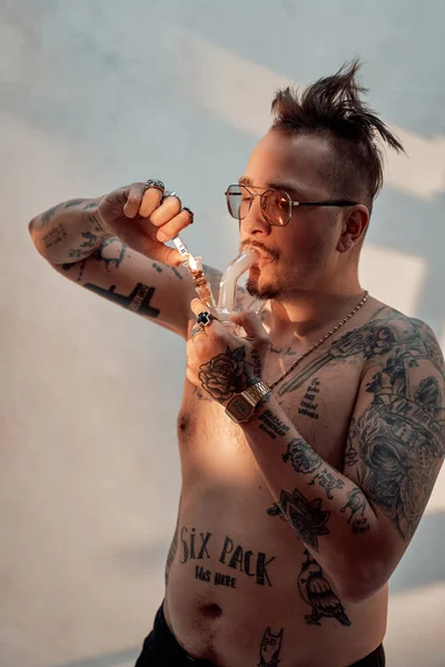 Shirtless man with tattooed body and freaky haircut lighting cannabis in the bong