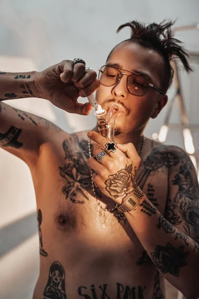 Hombre sin camisa con cuerpo tatuado y corte de pelo raro encendiendo cannabis en la pipa — Foto de Stock
