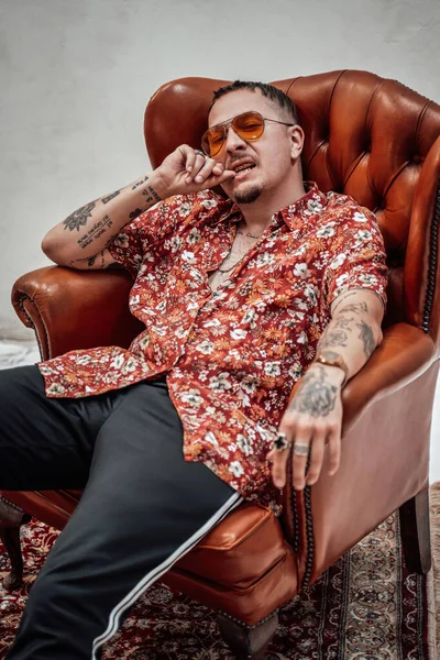 Tipo muy expresivo y elegante en camisa hawaiana y pantalones deportivos se sienta en sillón retro, se ve raro y loco — Foto de Stock