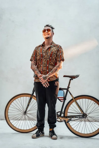 Elegante gángster tatuado freak en camisa hawaiana y pantalones deportivos de pie junto a una bicicleta hecha a medida, mira a la cámara con una sonrisa loca — Foto de Stock