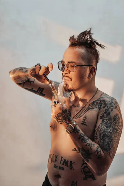 Homem sem camisa com corpo tatuado e cabelo estranho iluminando maconha no cachimbo — Fotografia de Stock