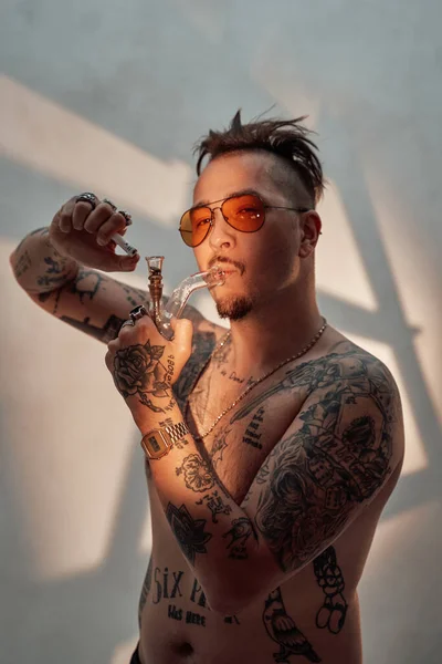 Homem sem camisa com corpo tatuado e cabelo estranho iluminando maconha no cachimbo — Fotografia de Stock
