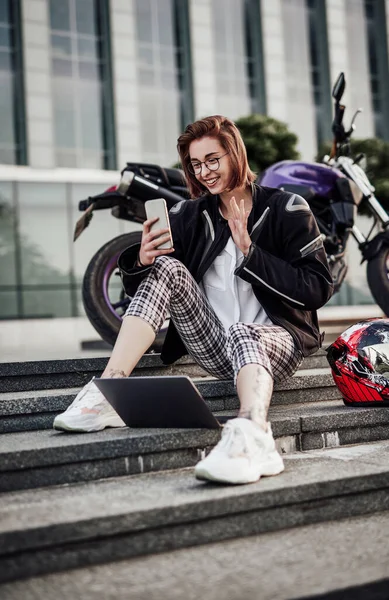 Glimlachend meisje zit in de buurt van haar motor en kijkt naar het scherm van haar telefoon — Stockfoto