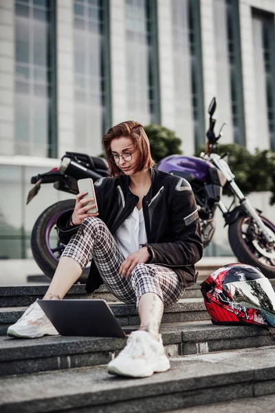 Jong meisje zit in de buurt van haar motor en kijkt naar het scherm van haar telefoon — Stockfoto