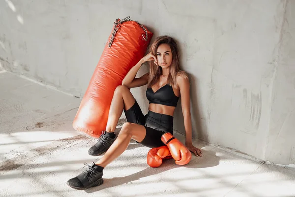 Sexy chica deportiva en un chándal negro con un saco de boxeo y guantes de boxeo —  Fotos de Stock