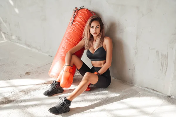 Chica bastante deportiva en un chándal negro con un saco de boxeo y guantes de boxeo — Foto de Stock