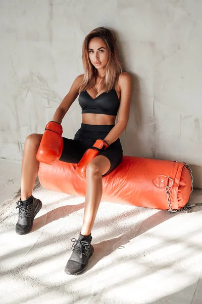 Chica de deportes calientes en un chándal negro con un saco de boxeo y guantes de boxeo — Foto de Stock
