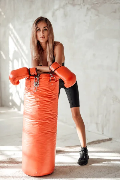Sexy chica deportiva, de pie en un chándal con un saco de boxeo y guantes de boxeo — Foto de Stock