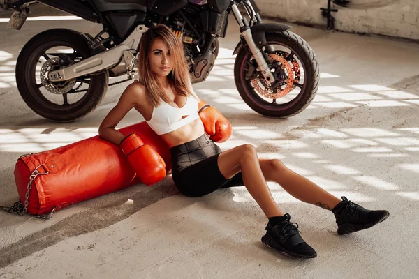 Hot and sexy sportswoman posing in a studio sitting on the floor with a punching bag. — Stock Photo, Image