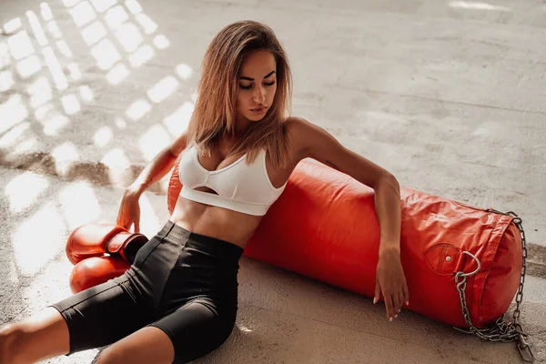 Attractive sportswoman sitting on the floor with a punching bag. — Stock Photo, Image