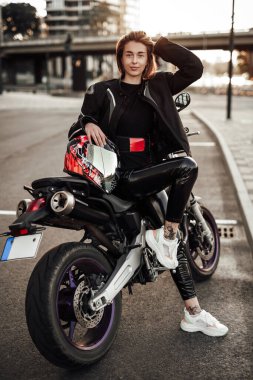Siyah ceketli, gülümseyen bir kız mor bir motosikletin üzerinde kaskıyla oturuyor.