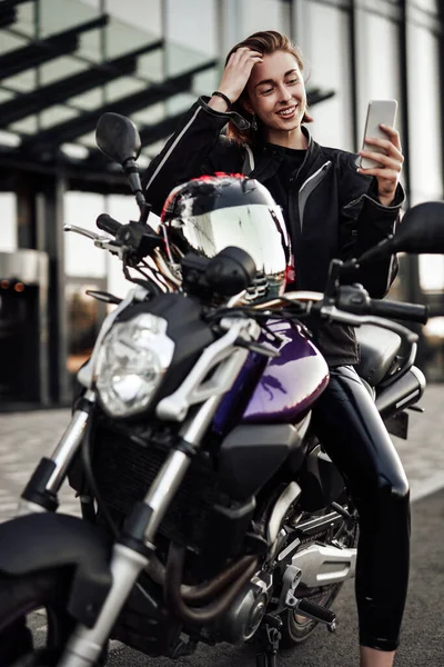 Jong meisje in een motorjack zit op een paarse motor en kijkt naar haar telefoon — Stockfoto