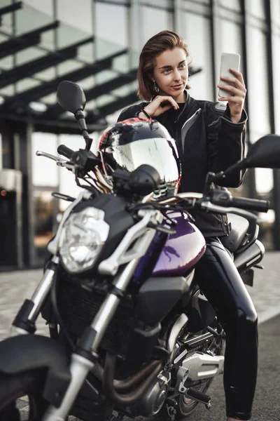 Κορίτσι σε ένα μπουφάν μοτοσικλέτας κάθεται σε ένα μοβ μοτοσικλέτα και κοιτάζει το τηλέφωνό της — Φωτογραφία Αρχείου