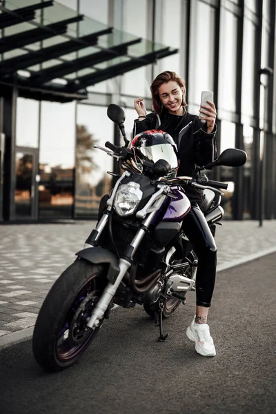 Ένα χαμογελαστό κορίτσι με ένα μπουφάν μοτοσικλέτας κάθεται σε ένα μοβ μηχανάκι και κοιτάζει το τηλέφωνό της — Φωτογραφία Αρχείου