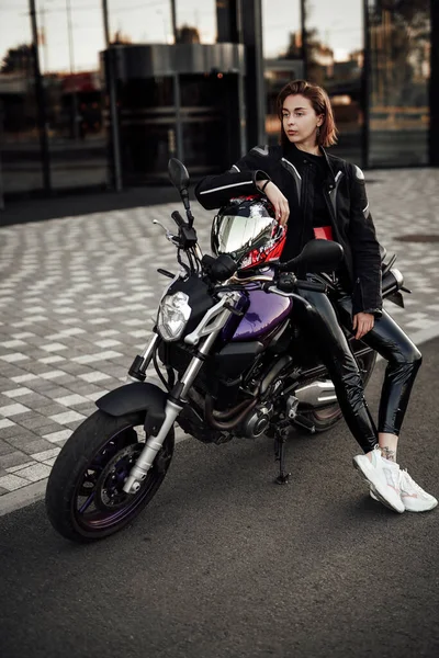 Menina sexy em uma jaqueta preta senta-se em uma moto roxa com um capacete de segurança vermelho — Fotografia de Stock