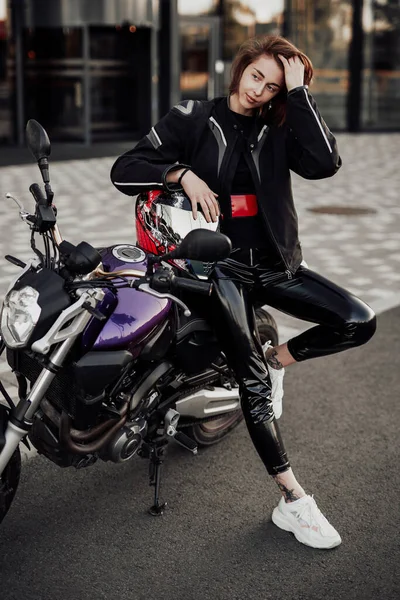Chica muy bonita en una chaqueta negra se sienta en una moto púrpura con un casco de seguridad rojo — Foto de Stock
