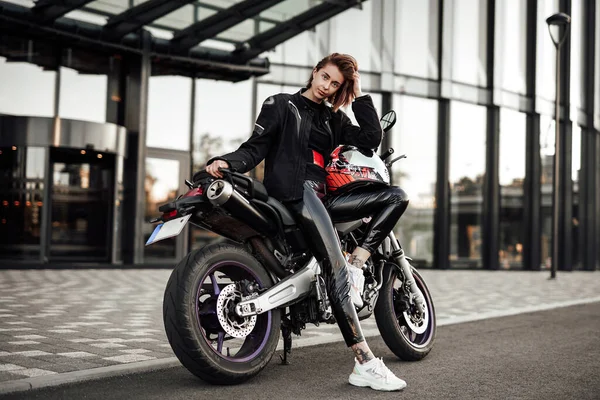 Menina quente em uma jaqueta preta senta-se em uma moto roxa com um capacete de segurança vermelho — Fotografia de Stock