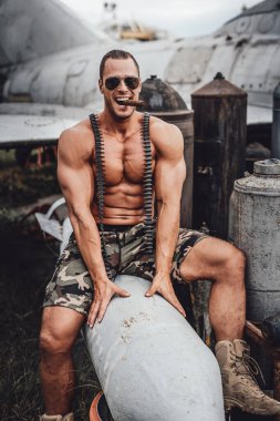 Amerikalı asker Küba purosu içiyor ve arka planda terk edilmiş bir uçakla oturuyor. Vahşi ve kaslı erkek asker..