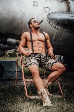 Terk edilmiş bir uçağın yanında purolu çıplak bir adam oturuyor.