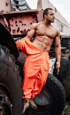 Elinde ingiliz anahtarı ve arkasında terk edilmiş bir makine olan çıplak bir adam.