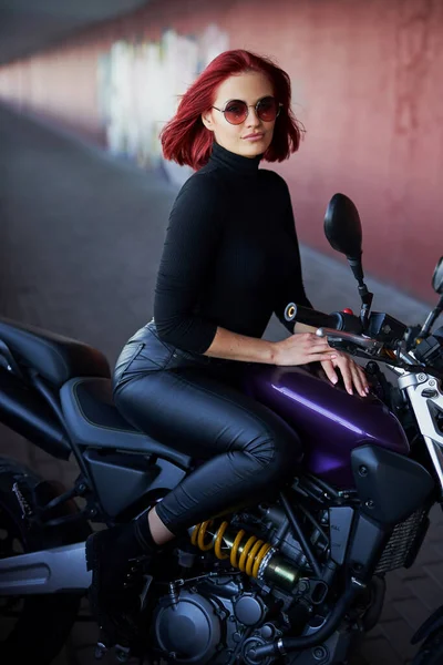 Uma menina com cabelos vermelhos posando com motocicleta urbana escura — Fotografia de Stock