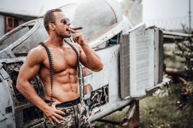 Güçlü bronzlaşmış adam arkasında uçakla puro içiyor.