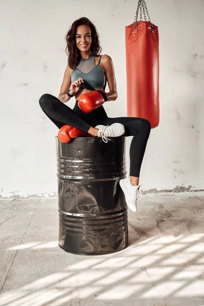 Slim woman with gloves sitting on barrel with bag behind — Stock Photo, Image