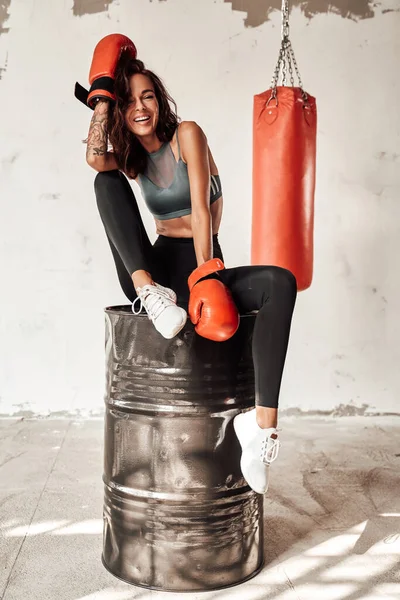 Slim woman with gloves sitting on barrel with bag behind — Stock Photo, Image
