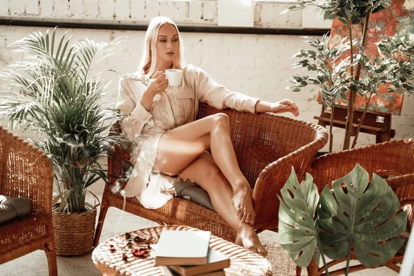 Chica glamour con taza de té posando en el sofá en la habitación acogedora — Foto de Stock