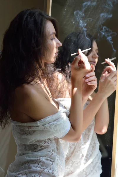 Sexy woman looking to the mirror and smoking at the window — Stock Photo, Image