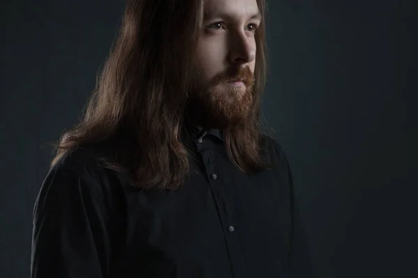 Portret van man met lange haren en baard in zwarte kleding op donkere achtergrond — Stockfoto
