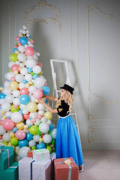 Een mooie blonde in een carnaval kostuum siert de boom van ballonnen. Charmante jongedame in een bochtige blauwe rok. — Stockfoto