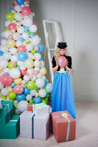 Söt och vacker blondin nära julgranen av ballonger och presenter i lådor. Charmig ung kvinna i en kurvig blå kjol. — Stockfoto