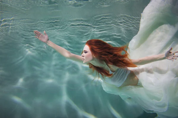 Underwater girl. Beautiful red-haired woman in a white dress, swimming under water. — Stock Photo, Image