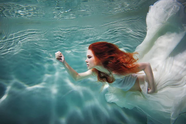 Underwater girl. Beautiful red-haired woman in a white dress, swimming under water. — Stock Photo, Image