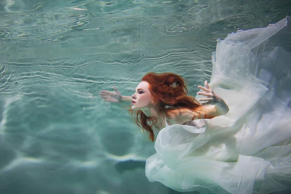 Underwater girl. Vacker rödhårig kvinna i en vit klänning, simma under vatten. — Stockfoto