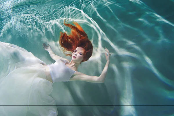 Underwater girl. Beautiful red-haired woman in a white dress, swimming under water. — Stock Photo, Image