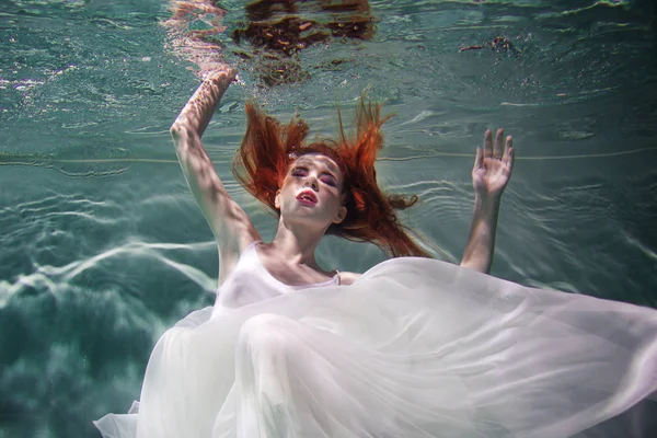 Underwater girl. Beautiful red-haired woman in a white dress, swimming under water. — Stock Photo, Image