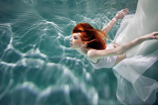 Underwater girl. Beautiful red-haired woman in a white dress, swimming under water. — Stock Photo, Image