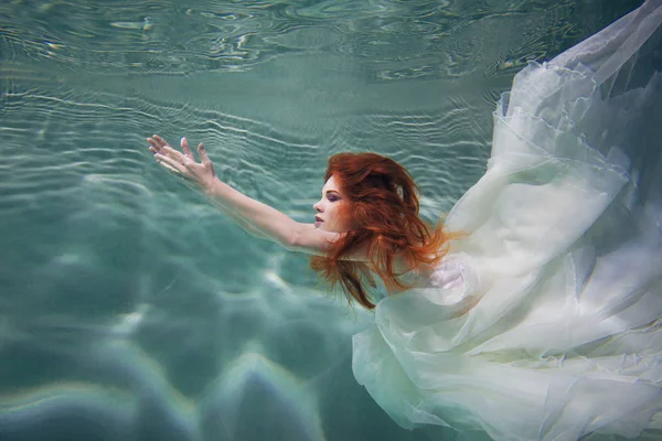 A víz alatti lány. Gyönyörű vörös hajú nő fehér ruhában, víz alatti úszás. — Stock Fotó
