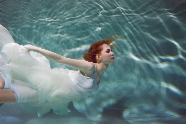 Underwater girl. Beautiful red-haired woman in a white dress, swimming under water. — Stock Photo, Image