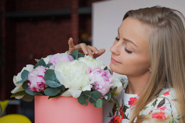 Cute young blonde woman sniffs a bouquet of flowers. Delicate pink peonies in a hat box